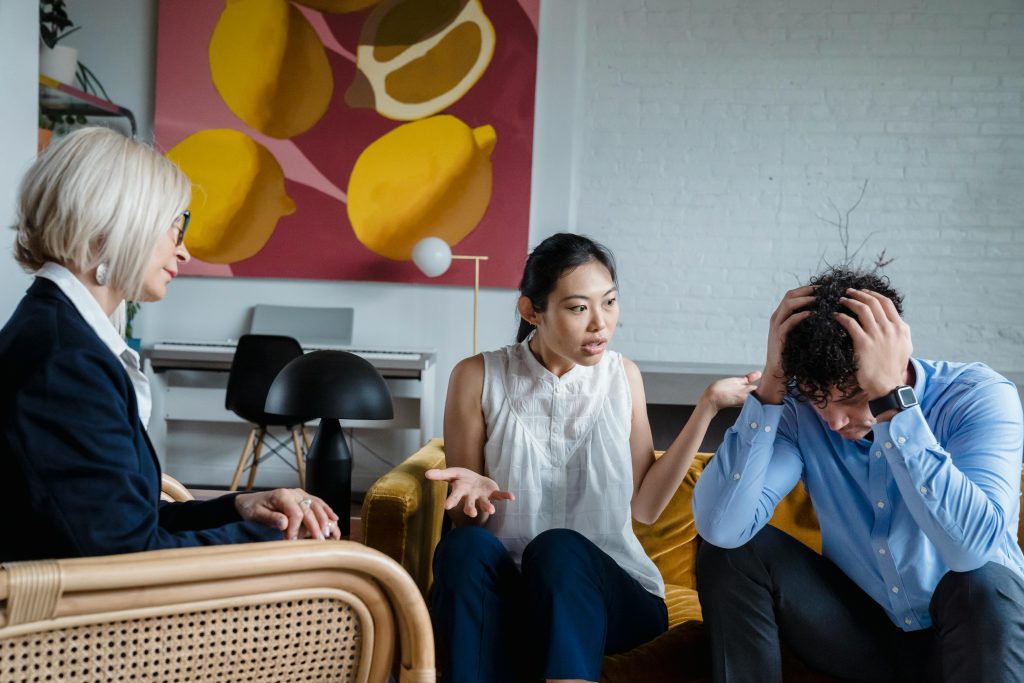 A young couple in a tense discussion sits on a yellow sofa while a woman in a blazer listens from a chair.